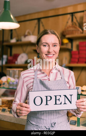 Smiling cheerful businesswoman wearing striped apron opening bakery Stock Photo