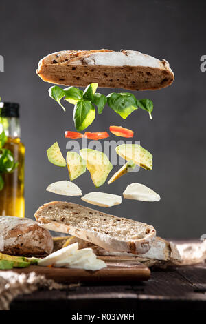 Isolated ingredients for a sandwich floating in air in layers with a crispy ciabatta bread, fresh basil, tomatoes, mozzarella and avocado over rustic  Stock Photo