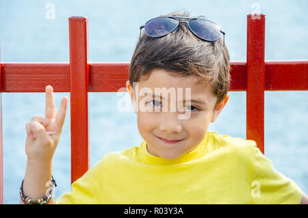 A young boy poses for a portrait and makes a peace sign with his fingers, wearing a yellow t-shirt with sunglasses on his head. Stock Photo