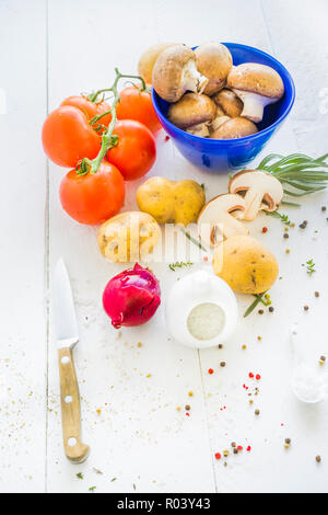 white tabletop displaying brown portobello mushrooms in a blue bowl, tomatoes, potatoes, a red onion, herbs and coarse sea salt and red, green Stock Photo