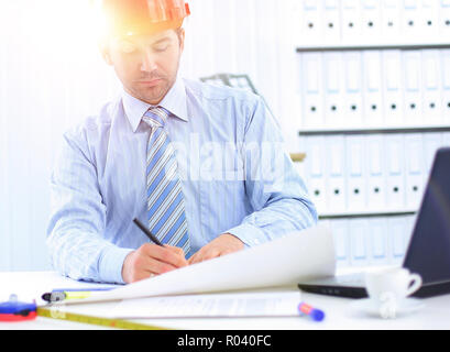 Architect looking working in office at desk. Wearing hardhat and taking notes on paper Stock Photo