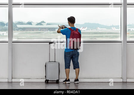 Malaysia, KLIA 2, 2018-04-03: Asian young man at the airport, uses mobile phone by the window. Tourist with a backpack and a suitcase, shoots a phone  Stock Photo
