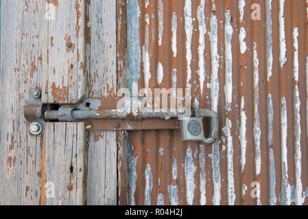 A rusty painted slide bolt secured with a Lockwood padlock on an old wooden and galvanized steel door with peeling red paint Stock Photo