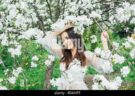 Portrait of young girl in white dress in blossom apple garden, touching tree branch Stock Photo
