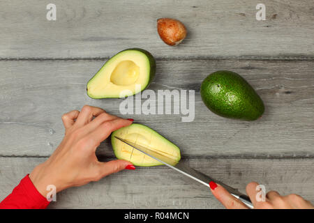 Tabletop view, woman hands holding chef knife and avocado cut in half, seed removed, cutting lines in pulp to portion it. Stock Photo