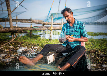 Fisherman mending net with traditional Chinese fishing nets behind, Fort Kochi (Cochin), Kerala, India, Asia Stock Photo