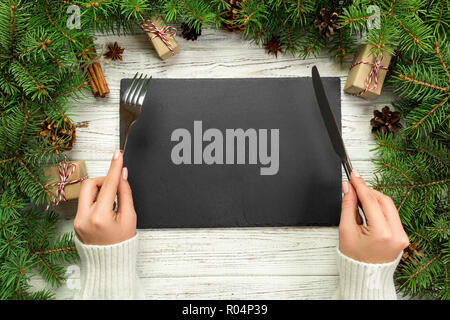 top view girl holds fork and knife in hand and is ready to eat. Empty black slate rectangular plate on wooden christmas background. holiday dinner dis Stock Photo