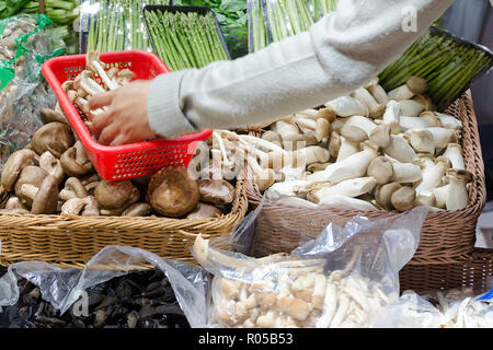 Stall with mushrooms and asparagus sprouts, hand fills the basket with mushrooms. Photo with blur in motion. Hong Kong, China. Stock Photo