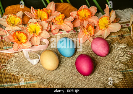 Painted Easter eggs and large pink daffodils with a yellow fringe in the middle, biscuits and burlap lie on a straw carpet Stock Photo