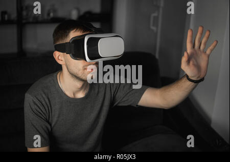 Young bearded man wearing headset of virtual reality in office sitting on a sofa and trying to touch something that he see in virtual world. Stock Photo