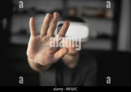 Young bearded man wearing headset of virtual reality in office sitting on a sofa and trying to touch something that he see in virtual world. Stock Photo