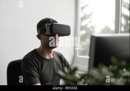 Young man wearing virtual reality goggles headset, vr box and sitting in the office against computer monitor. Connection, technology, new generation, progress concept. Stock Photo