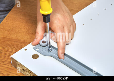The handyman tightens the screw in drawer runner of wooden table with a screwdriver, flat pack furniture assembly. Stock Photo