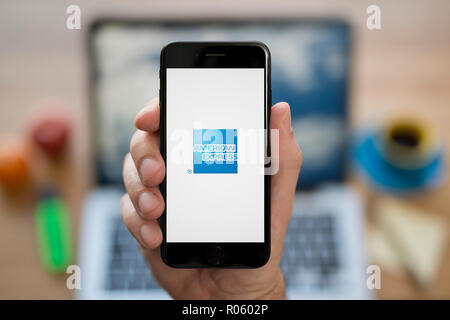 A man looks at his iPhone which displays the American Express logo, while sat at his computer desk (Editorial use only). Stock Photo