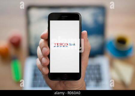 A man looks at his iPhone which displays the Tesco Bank logo, while sat at his computer desk (Editorial use only). Stock Photo