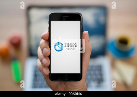 A man looks at his iPhone which displays the Tesco Mobile logo, while sat at his computer desk (Editorial use only). Stock Photo