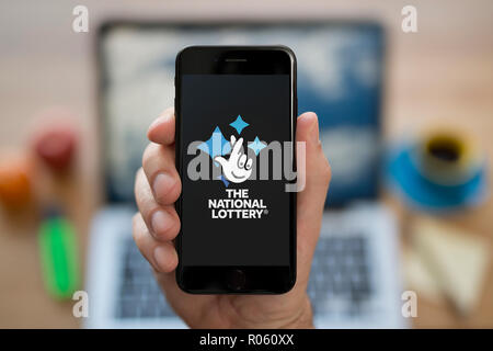 A man looks at his iPhone which displays the National Lottery logo, while sat at his computer desk (Editorial use only). Stock Photo