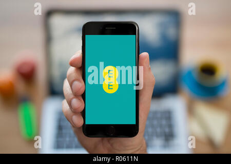 A man looks at his iPhone which displays the EE logo, while sat at his computer desk (Editorial use only). Stock Photo