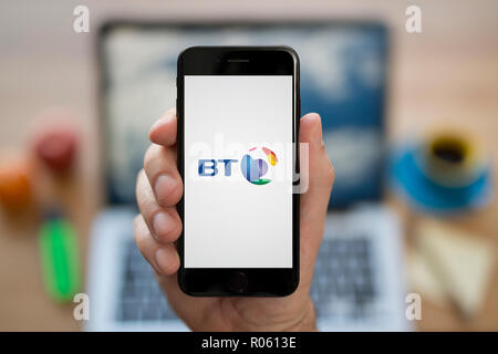 A man looks at his iPhone which displays the BT logo, while sat at his computer desk (Editorial use only). Stock Photo