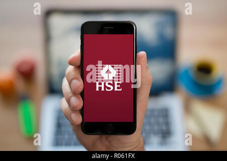 A man looks at his iPhone which displays the Health and Safety Executive (HSE) logo, while sat at his computer desk (Editorial use only). Stock Photo