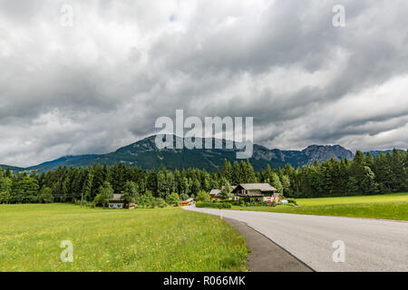 Alps in Austria. Wooden house before mountains green field and road through green pine forest Stock Photo