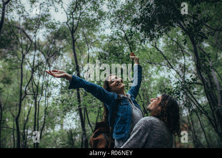 Young woman stretching out arms while carried by her boyfriend. Dating couple enjoying the rain in forest. Stock Photo