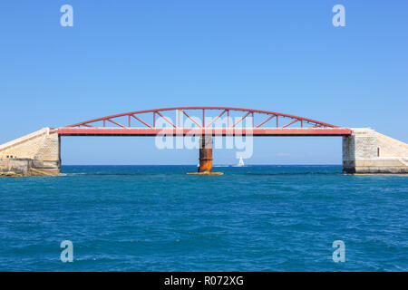 Small white sailboat ship in blue water of Grand Harbor, Valletta. Red Breakwater Bridge metallic construction on foreground in sunny day Stock Photo