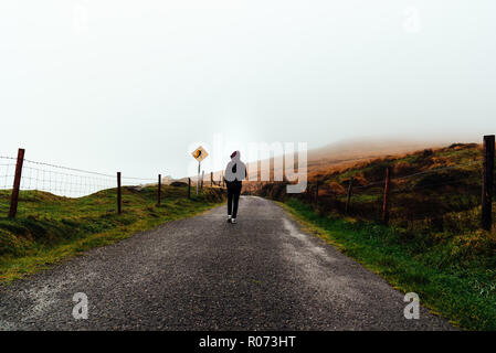 Young woman on hoodie walks alone on misty road. Stock Photo