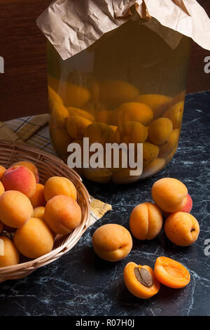 Ripe organic apricots harvested at summer time and big glass jar with canned sweet apricot compote on dark marble. Composition in rustic style - organ Stock Photo