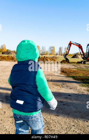 Back view on cute little toddler boy standing on playground and looking at big construction tractor, digging land in rural. Stock Photo