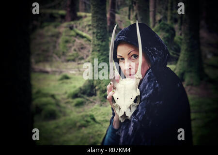 Female witch standing alone in dark woods holding a deer skull - John Gollop Stock Photo