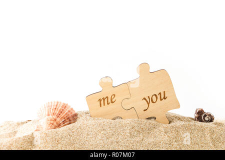 Two pieces of wooden puzzle with Me & You words, on sand with seashells and white background. Stock Photo