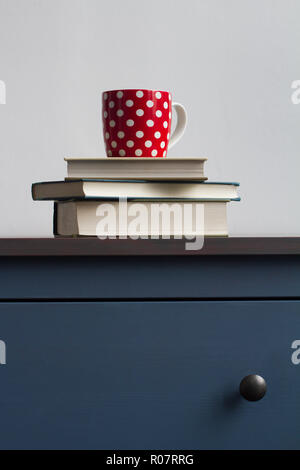 vertical front view detail of stack closed books with red dotted tea cup on blue vintage wooden chest of drawers with black handles against wall in mo