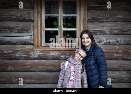 Mum and daughter embracing against the background of an old wooden log wall with a window. Stock Photo