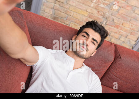 Portrait of a serious handsome young man talking a selfie while sitting on a sofa in a white tshirt from the perspective of the smartphone. Stock Photo