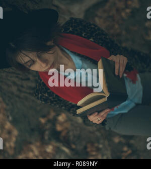 Young woman with book in her hands sits next to tree trunk and reads during of dusk. Top view. Stock Photo