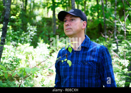 A caucasian man is looking away while hiking in the forest wearing a blue check shirt and a black hat. Stock Photo