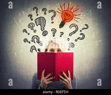 Young pensive woman hiding behind a red book interrogation marks, lightbulbs and arrow mess above head. Question and idea concept. Find solution to pr Stock Photo