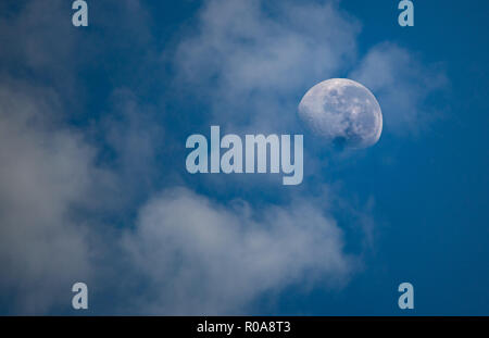 A daytime moon visible through soft, passing clouds against a clear blue sky. A beautiful and serene view of celestial details in daylight. Stock Photo