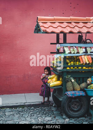 Small young Guatemalan girl in traditional Mayan dress hiding behind her family's fruit cart street stall Stock Photo