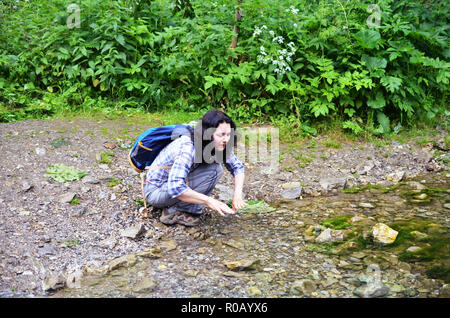 Traveling woman washing her face and hands with water from stream in forest. Girl sits with backpack Stock Photo