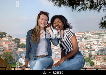 Two young female friends - one African, one European, sitting on a wall on a summer's day with the skyline of Lisbon behind them Stock Photo