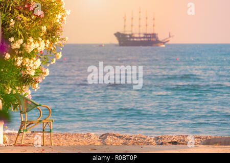 Beautiful scene with cruise sailboat sailing in sea at sunset at horizon. View from beach with chair in foreground. Stock Photo