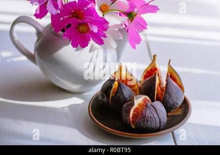 White vase with flowers. Ripe figs in a plate on a white wooden background. Sunny day. Stock Photo