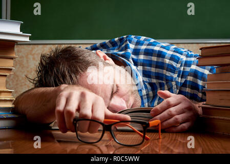 A tired and tortured disheveled student with glasses is sleeping at a table with stacks of books against the background of a cool blackboard. Stock Photo
