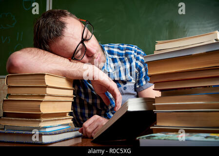 Tired and tortured disheveled student in glasses is sleeping at the table on a stack of books against the background of the chalkboard. Stock Photo