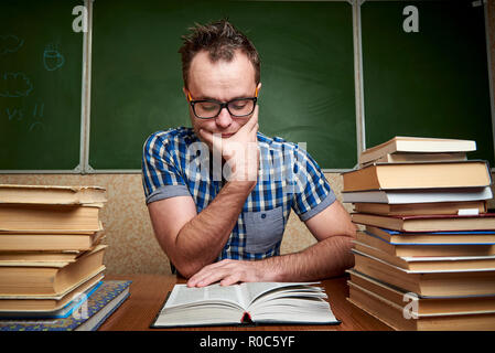 Disheveled unshaven young man in glasses reading a book at the table with piles of books on the background of the blackboard. Stock Photo