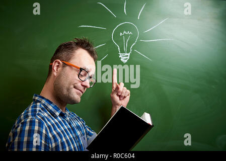 A young disheveled man with glasses is reading a book, an idea comes to mind on the background of a blackboard with a chalked light bulb. Stock Photo