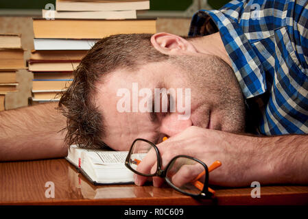 A tired and tortured disheveled student with glasses is sleeping at a table with stacks of books against the background of a cool blackboard. Stock Photo