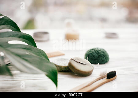 Eco friendly natural bamboo toothbrushes, shampoo bar, toothpaste in glass, wooden brush and konjaku sponge on white wood with green monstera leaves.  Stock Photo